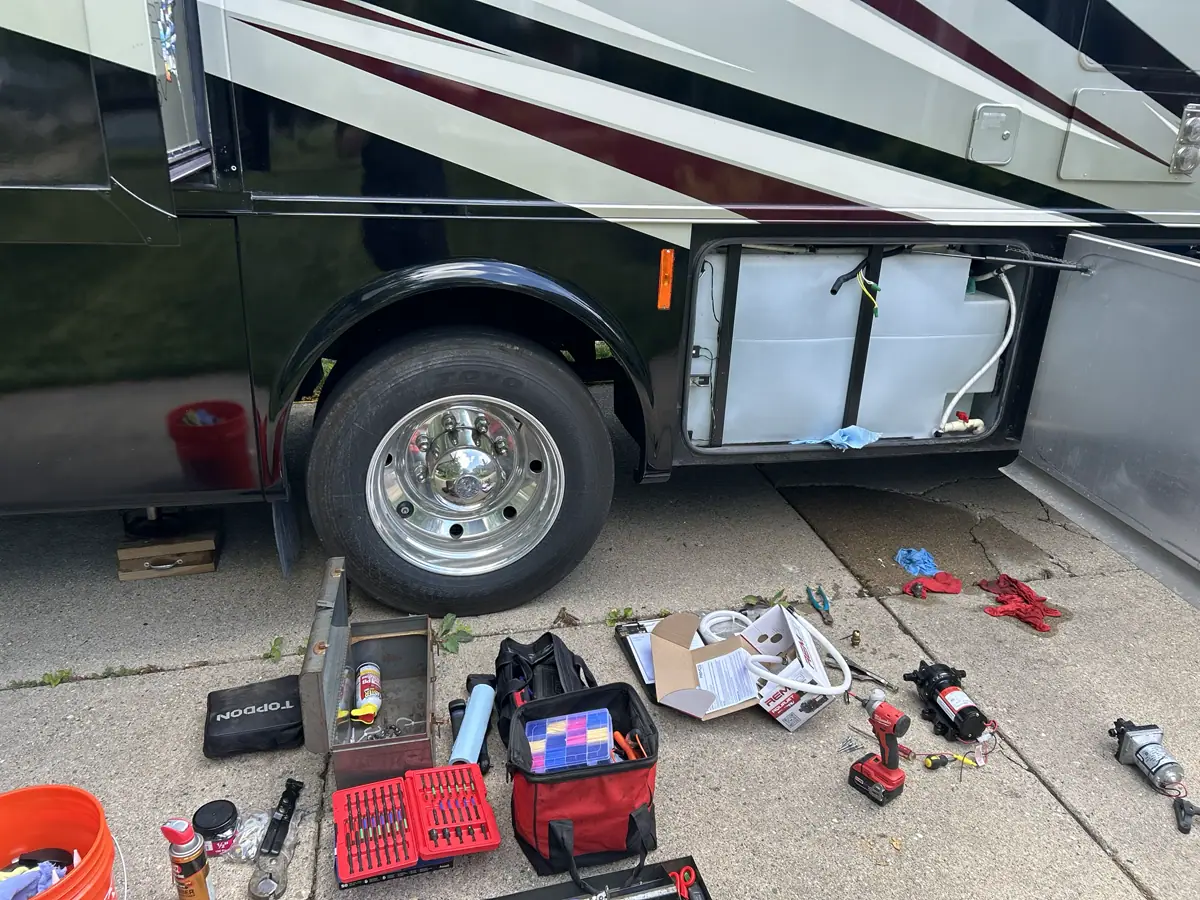 Tools and a new water pump beside an open RV fresh-water tank compartment during a plumbing repair.